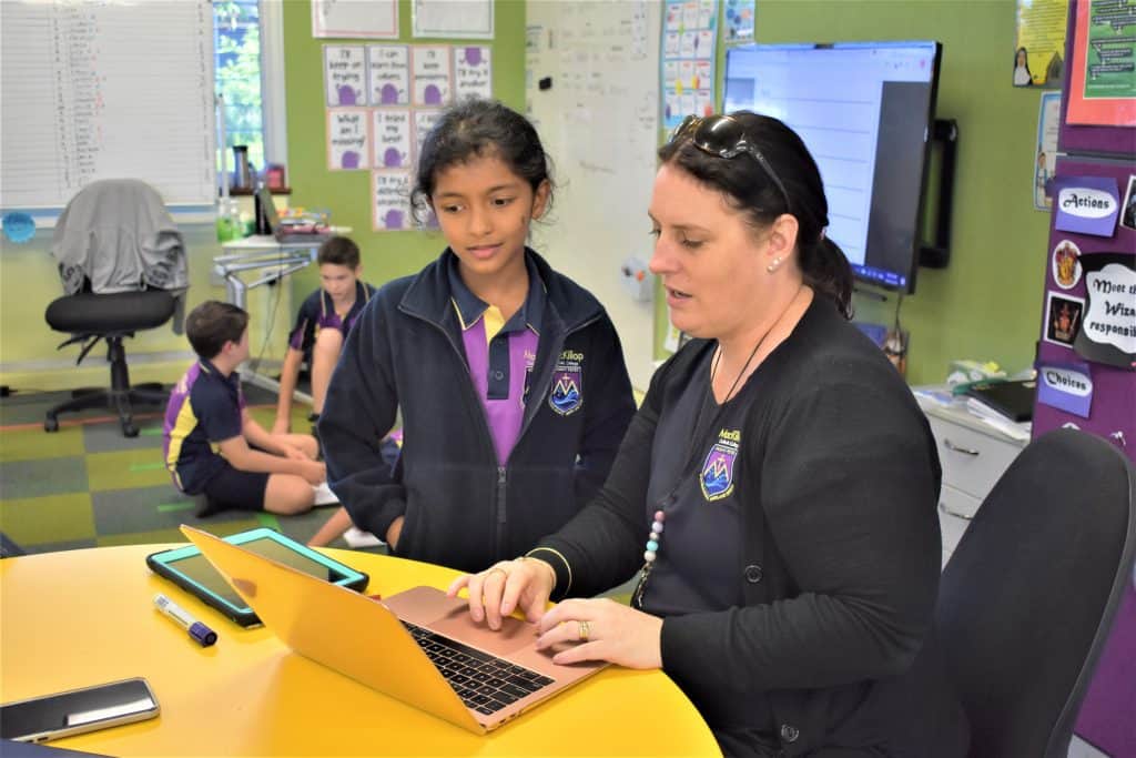 Students at MacKillop Catholic College,, an outstanding school in Cairns, recieves help from teacher in classroom. Other students are int he background. 